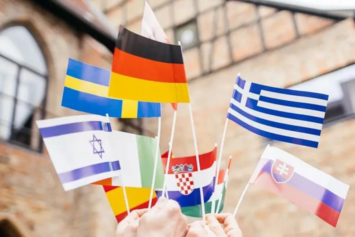 Mehrere Hände halten kleine internationale Flaggen in die Höhe, darunter unter anderem die Flaggen von Deutschland, Griechenland, Schweden, Israel, Kroatien, der Slowakei, Italien und Litauen. Im Hintergrund ist ein historisches Gebäude zu sehen. Das Bild symbolisiert internationale Vielfalt und kulturellen Austausch an der Universität Liechtenstein.