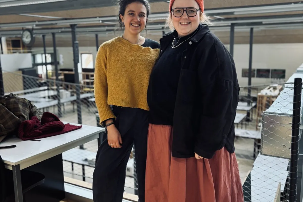 Zwei Personen stehen lächelnd in einem modernen Architekturatelier mit großen Fenstern und industriellem Design. Sie tragen lässige, kreative Kleidung. Im Hintergrund sind Arbeitsplätze und eine Uhr sichtbar.