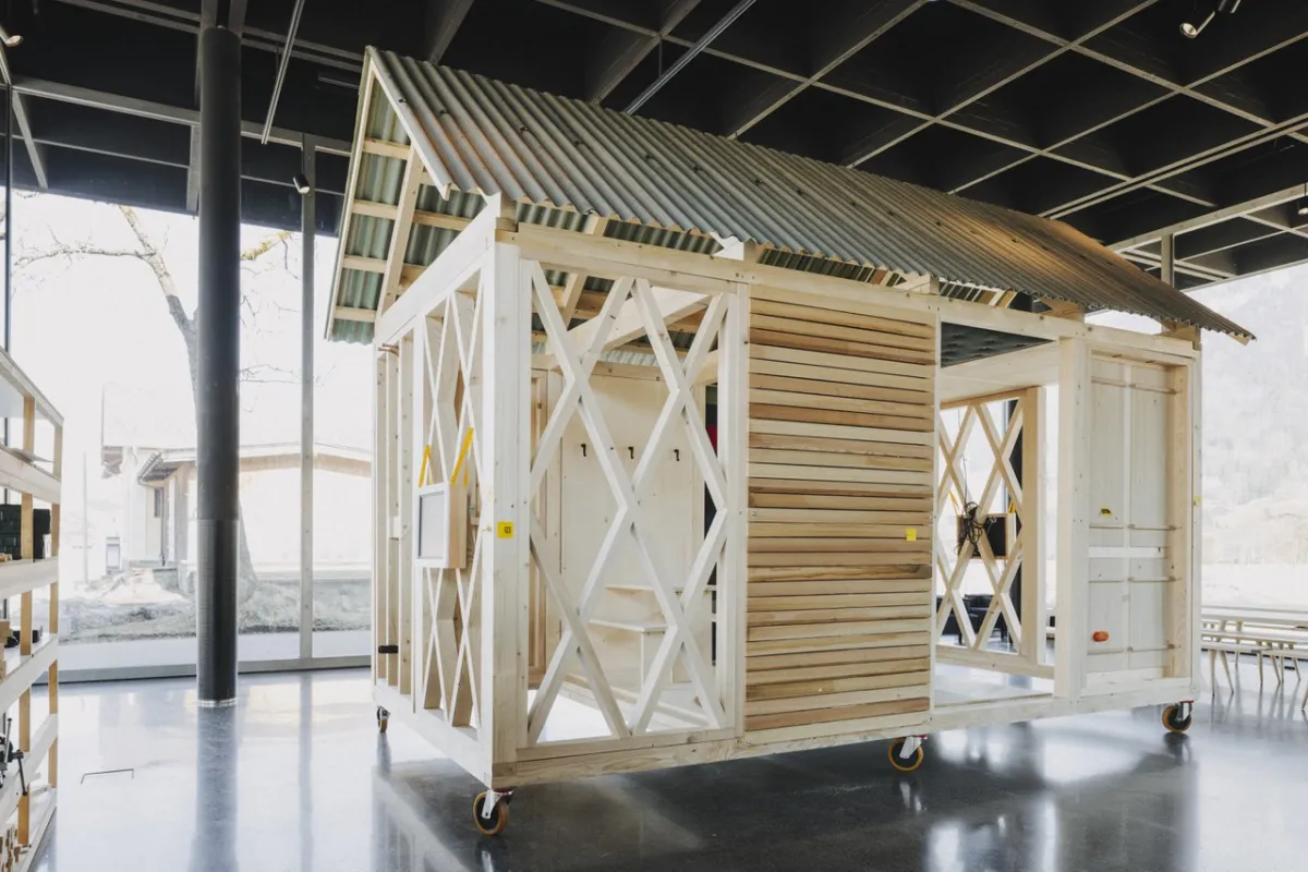 Ein einfaches, modulares Holzgebäude mit einem Wellblechdach steht auf Rollen in einem modernen, lichtdurchfluteten Raum. Das Design zeigt eine offene Struktur mit diagonalen Verstrebungen und Holzverkleidung, ideal für nachhaltiges Bauen und architektonische Bildung.