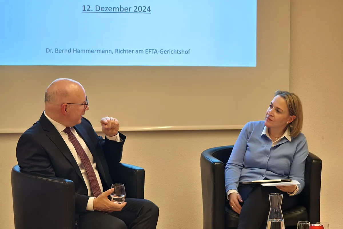 Zwei Personen sitzen in Sesseln vor einer Leinwand, die das Datum "12. Dezember 2024" zeigt. Eine Person hält ein Glas Wasser, die andere hat ein Notizbuch auf dem Schoß. Im Vordergrund steht eine Karaffe auf einem Tisch mit weihnachtlicher Dekoration.