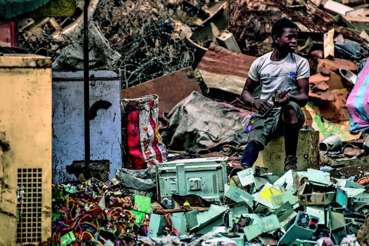 Ein Mann sitzt auf einem Hocker inmitten von Elektroschrott auf einer Müllhalde. Um ihn herum sind alte Geräte und Kabel zu sehen. Die Szene zeigt die Problematik von Elektroschrott und dessen Auswirkungen auf die Umwelt.