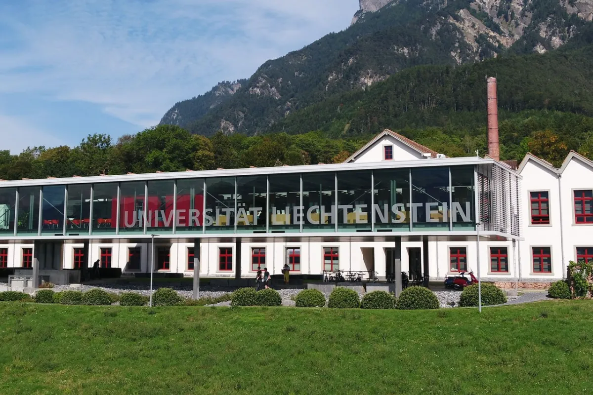 Außenansicht der Universität Liechtenstein mit modernem Glasgebäude und traditionellem weißen Gebäude im Hintergrund, umgeben von grüner Landschaft und Bergen.