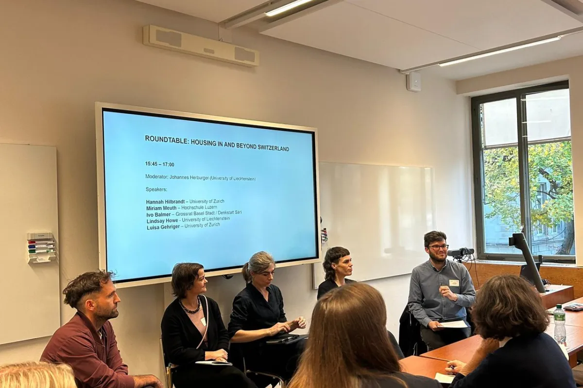 Eine Gruppe von Personen sitzt in einem Seminarraum und diskutiert bei einer Podiumsdiskussion zum Thema "Housing in and Beyond Switzerland". Im Hintergrund ist ein Bildschirm mit den Namen der Teilnehmenden und dem Moderator zu sehen.