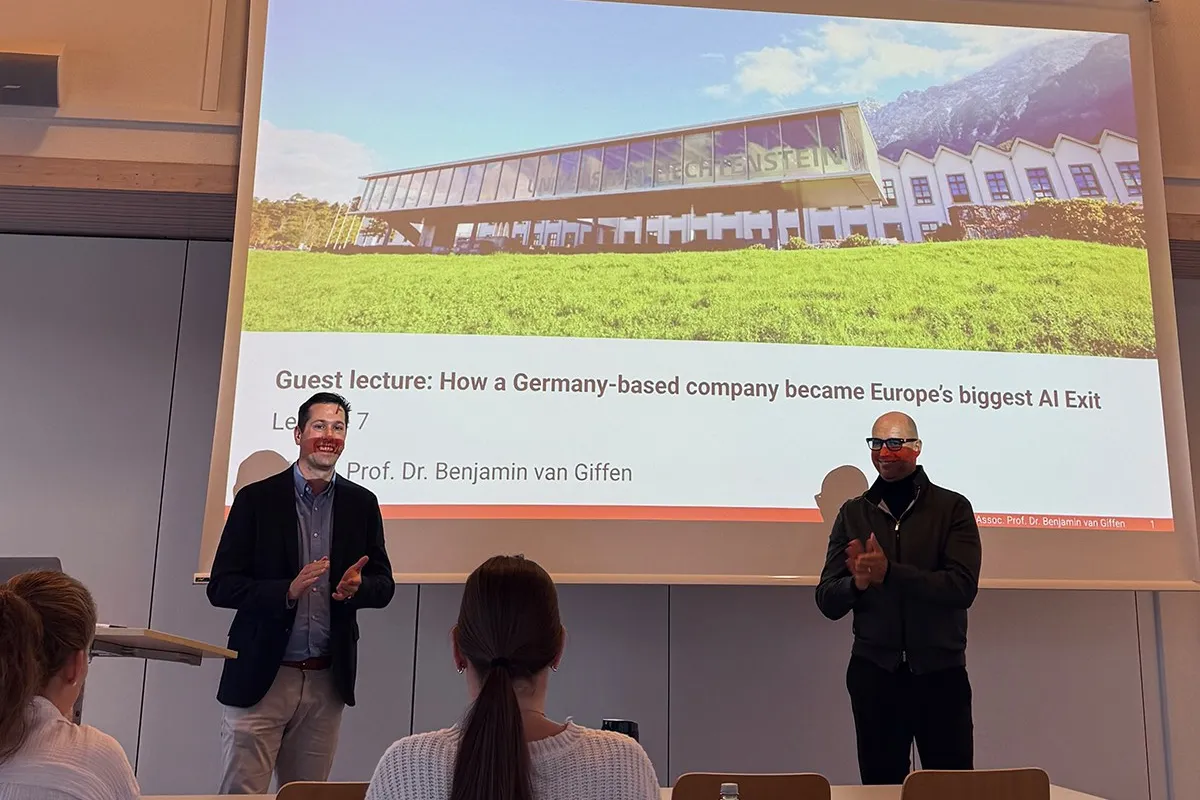 Zwei Männer stehen vor einer Präsentationsfolie in einem Vorlesungssaal der Universität Liechtenstein. Die Folie zeigt den Titel eines Gastvortrags über den Erfolg eines deutschen KI-Unternehmens. Studierende sitzen im Publikum.
