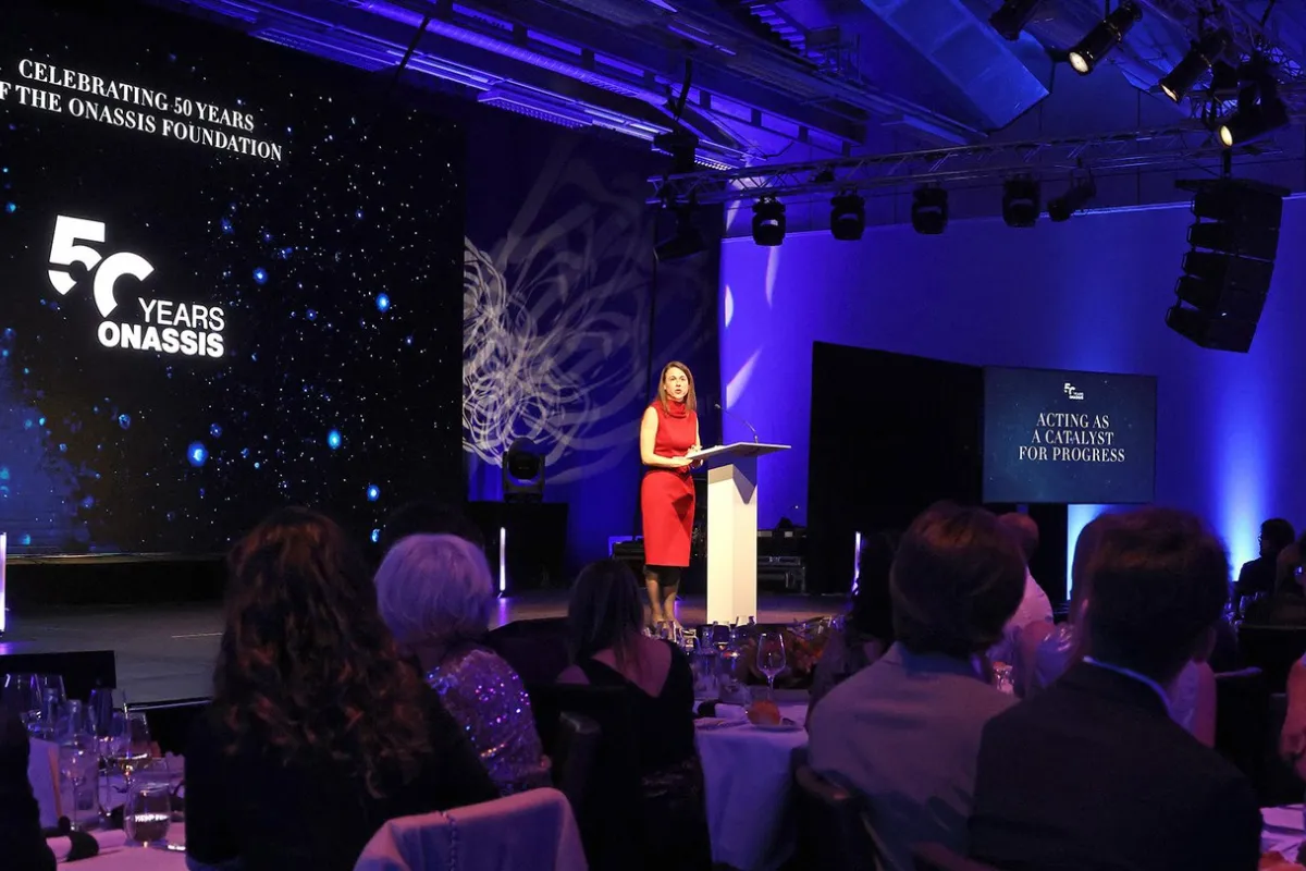 Eine Rednerin in rotem Kleid steht an einem Podium auf einer Bühne, umgeben von Publikum. Im Hintergrund ist eine Leinwand mit der Aufschrift "Celebrating 50 Years of the Onassis Foundation" zu sehen. Die Veranstaltung findet in einem festlich beleuchteten Raum statt.