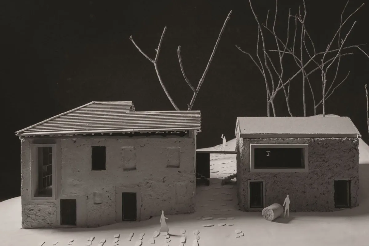 Modell eines architektonischen Projekts mit zwei Gebäuden, umgeben von kahlen Bäumen. Das linke Gebäude hat ein geneigtes Dach, während das rechte ein Flachdach aufweist. Kleine Figuren und Fußspuren sind auf dem Boden sichtbar, was eine menschliche Präsenz andeutet.