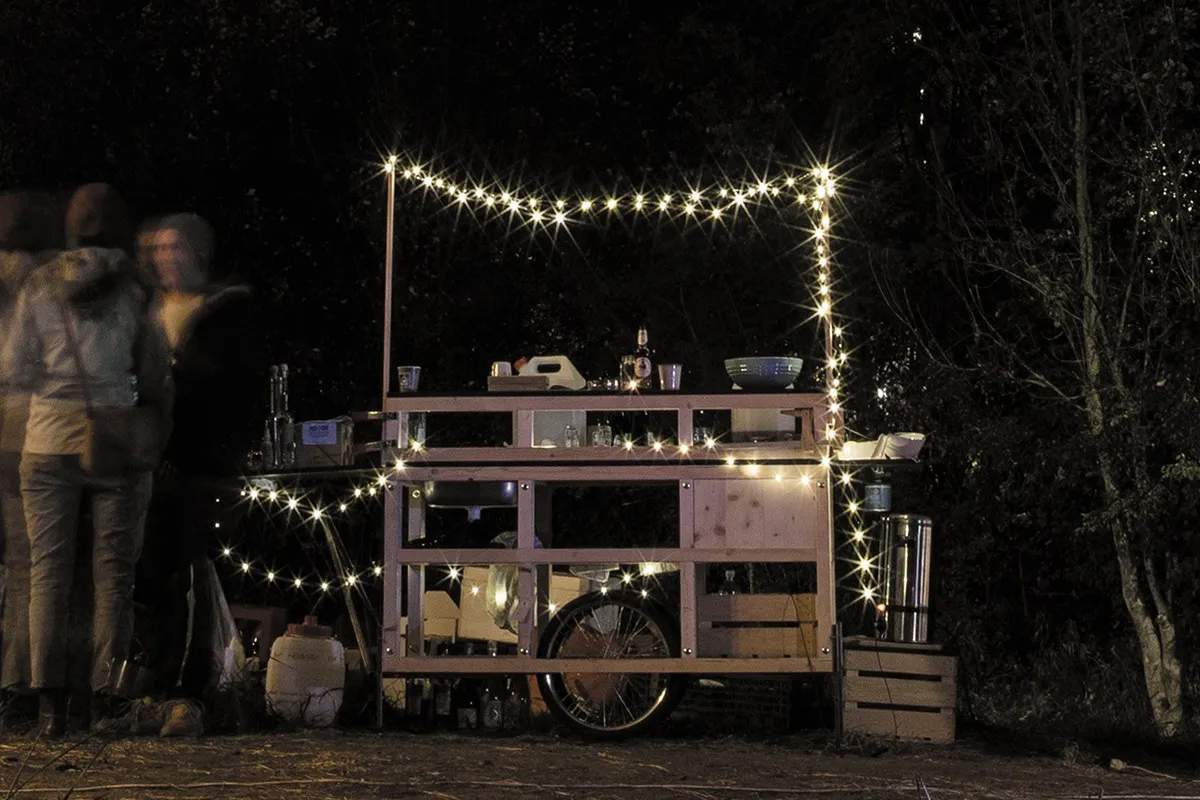 Eine mobile Holzbar, dekoriert mit Lichterketten, steht im Freien bei Nacht. Mehrere Personen stehen links neben der Bar und unterhalten sich. Die Szene vermittelt eine gemütliche und einladende Atmosphäre.