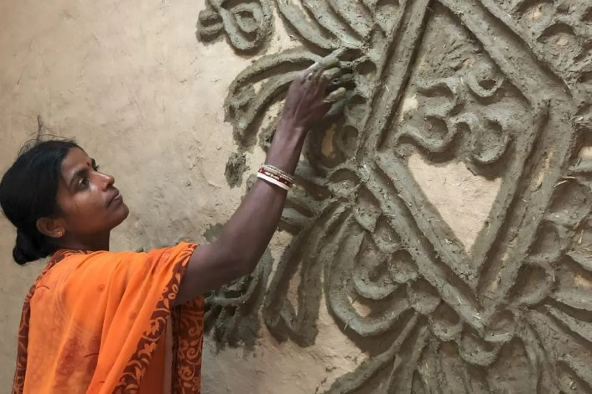Eine Frau in einem orangefarbenen Sari verziert eine Wand mit traditionellen Tonmustern. Sie arbeitet konzentriert an einem kunstvollen Relief.