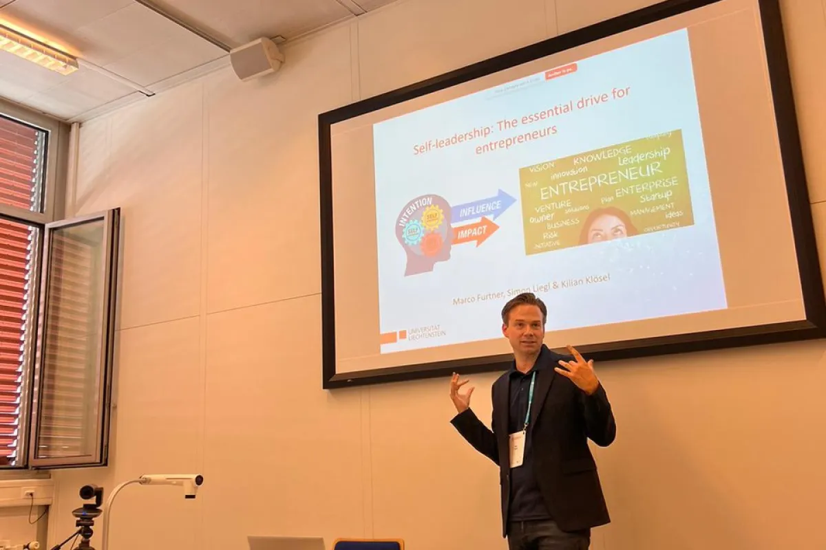 Ein Vortragender präsentiert in einem Seminarraum ein Paper über Selbstführung für Unternehmer. Im Hintergrund ist eine Leinwand mit einer Präsentationsfolie zu sehen. Tische mit Laptops und Kamera sind im Vordergrund.
