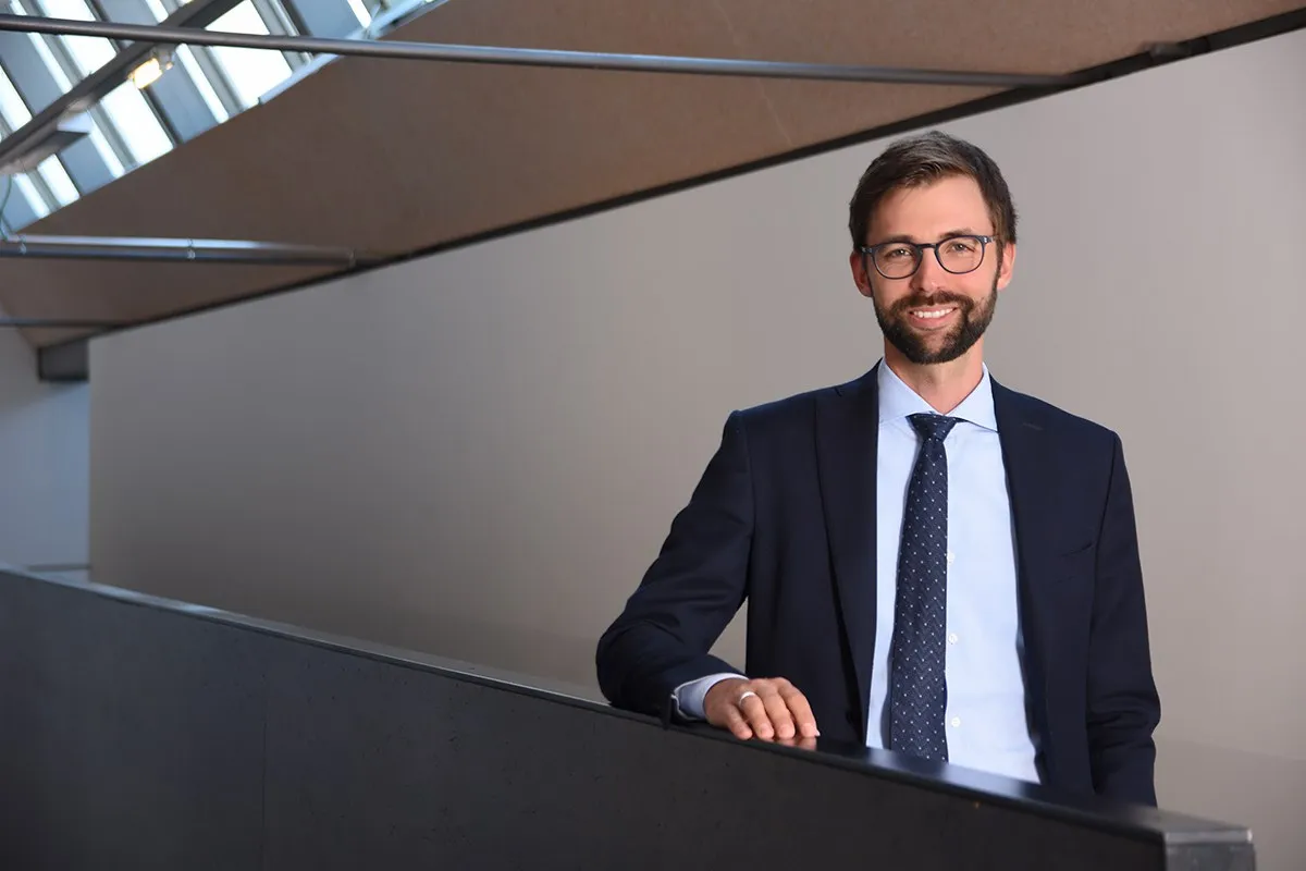 Ein Mann in Anzug und Krawatte steht lächelnd in einem modernen Gebäude mit schrägen Decken und großen Fenstern.