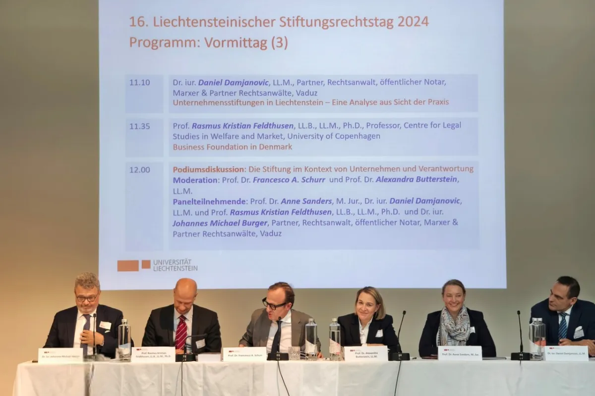 Podiumsdiskussion beim 16. Liechtensteinischen Stiftungsrechtstag 2024 an der Universität Liechtenstein. Fünf Expertinnen und Experten sitzen an einem Tisch vor einer Leinwand mit dem Tagesprogramm.