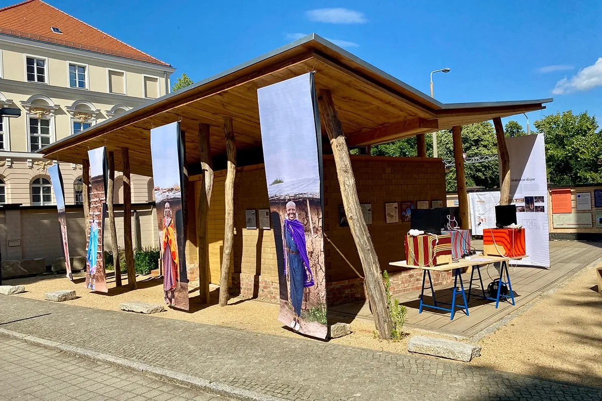 Ein hölzerner Pavillon mit großen, vertikalen Bannern, die Maasai-Frauen in traditioneller Kleidung zeigen. Im Vordergrund stehen Tische mit bunten Stoffen und Modellen. Der Pavillon ist Teil einer Ausstellung über Maasai-Baumeisterinnen und nachhaltige Architektur.