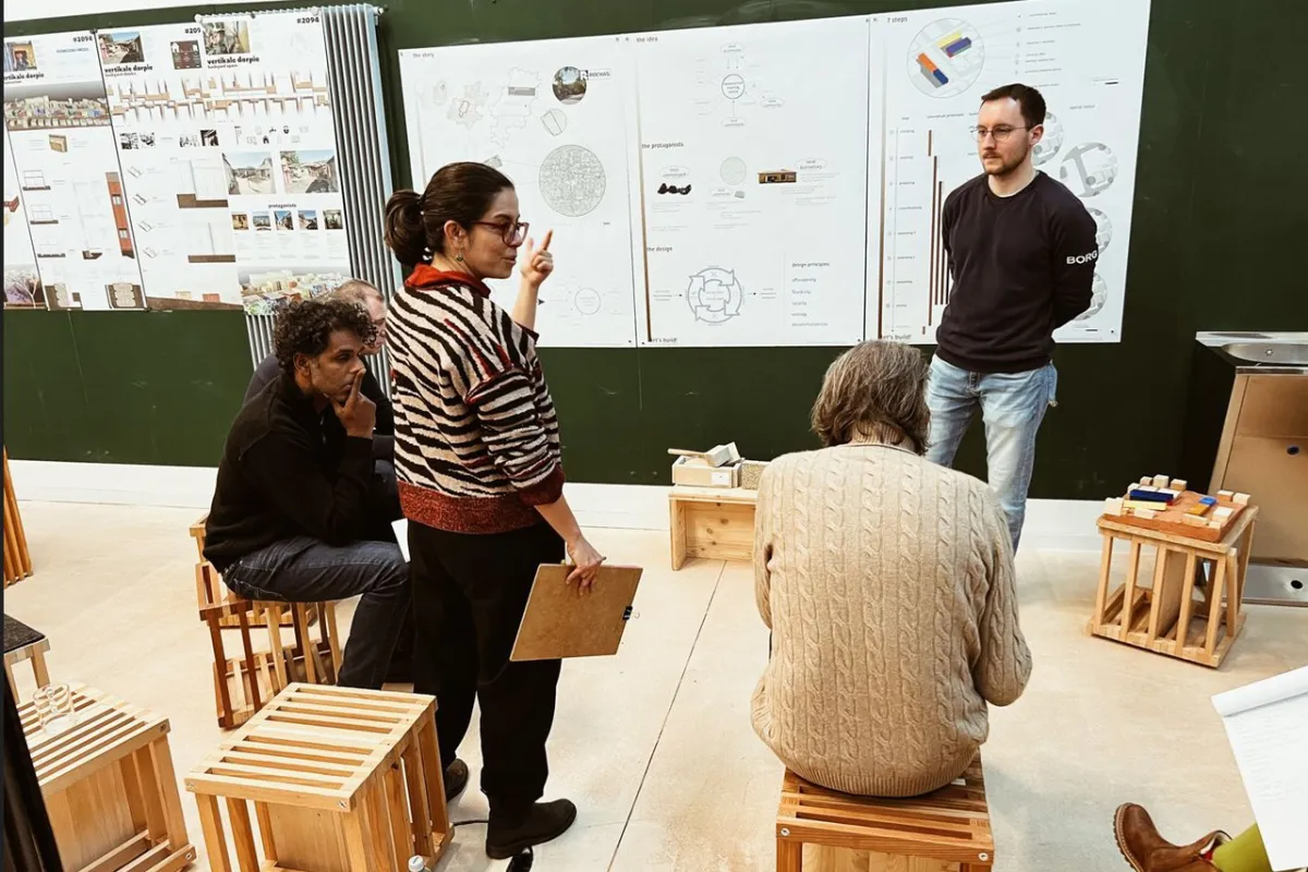 Eine Gruppe von Personen diskutiert vor einer Wand mit Architekturplänen. Sie sitzen auf Holzhockern in einem Seminarraum. Eine Frau steht und gestikuliert, während sie auf die Pläne zeigt.