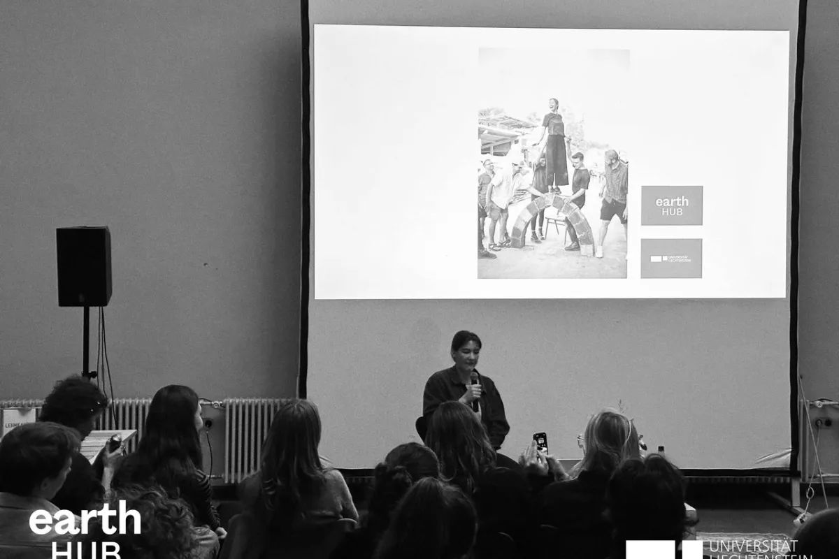 Eine Vortragende steht vor einem Publikum und präsentiert. Im Hintergrund ist eine Leinwand zu sehen. Das Bild ist in schwarz-weiss.