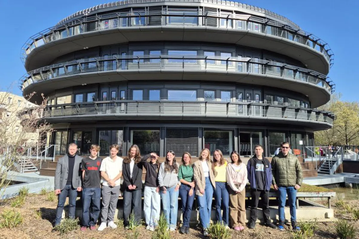 Gruppe von Studierenden und einer Lehrperson vor einem modernen, runden Universitätsgebäude bei sonnigem Wetter.