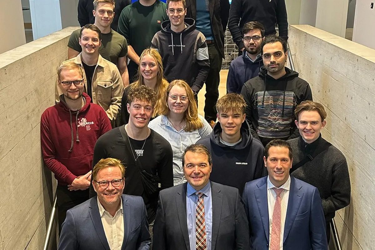 Gruppe von Studierenden und Lehrenden steht auf einer Treppe in einem modernen Gebäude der Universität Liechtenstein, lächelnd in die Kamera. Im Hintergrund sind Bildschirme und eine Steinwand zu sehen.