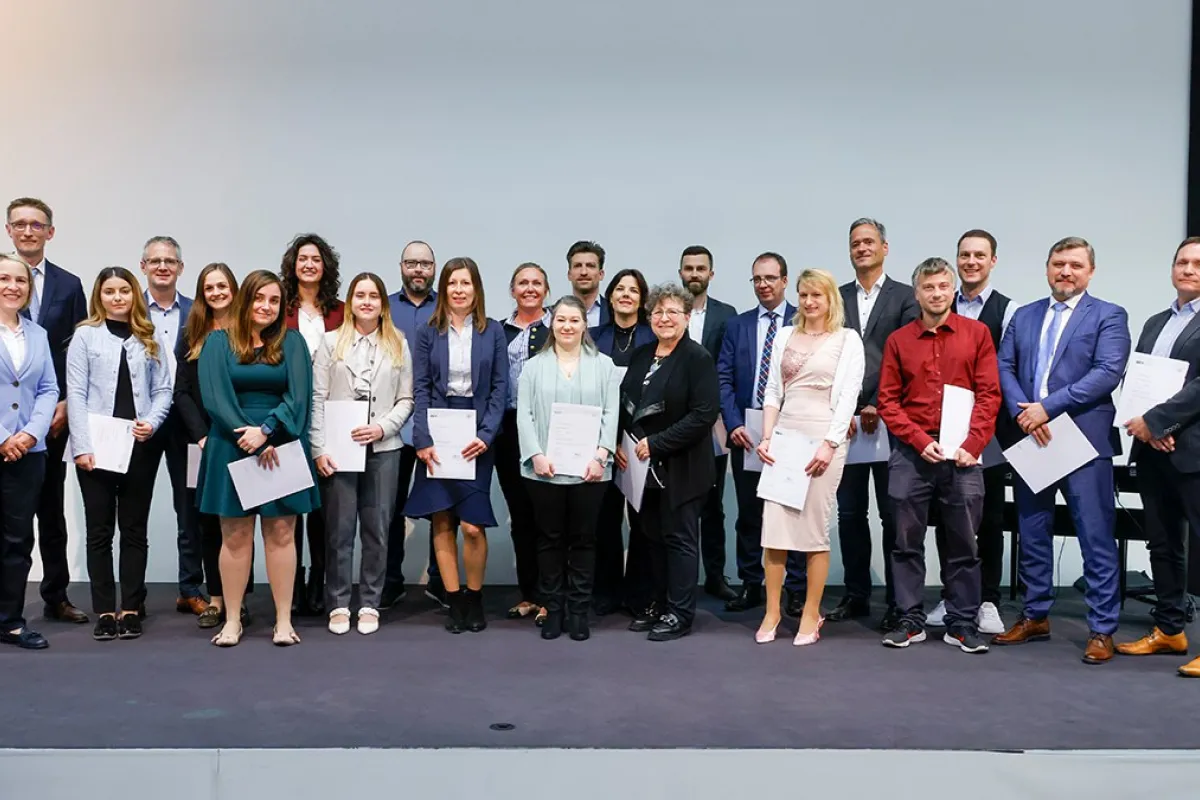 Eine Gruppe von Absolventinnen und Absolventen steht auf einer Bühne und hält Zertifikate in den Händen. Sie lächeln in die Kamera. Die Personen sind formell gekleidet, und im Hintergrund ist eine neutrale Wand zu sehen.