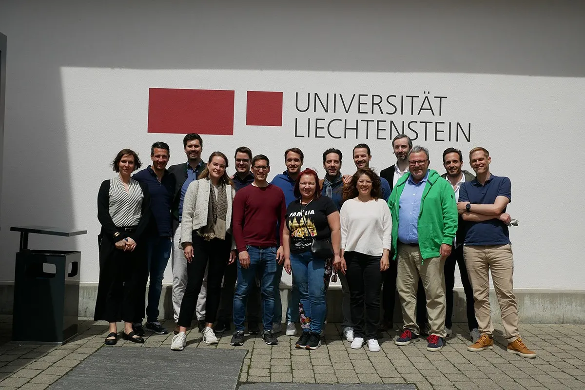 Gruppenfoto von Alumni des Abschlussjahrgangs 2013 des Masters in Banking and Financial Management vor dem Logo der Universität Liechtenstein. Die Personen stehen lächelnd zusammen auf dem Campus bei sonnigem Wetter.