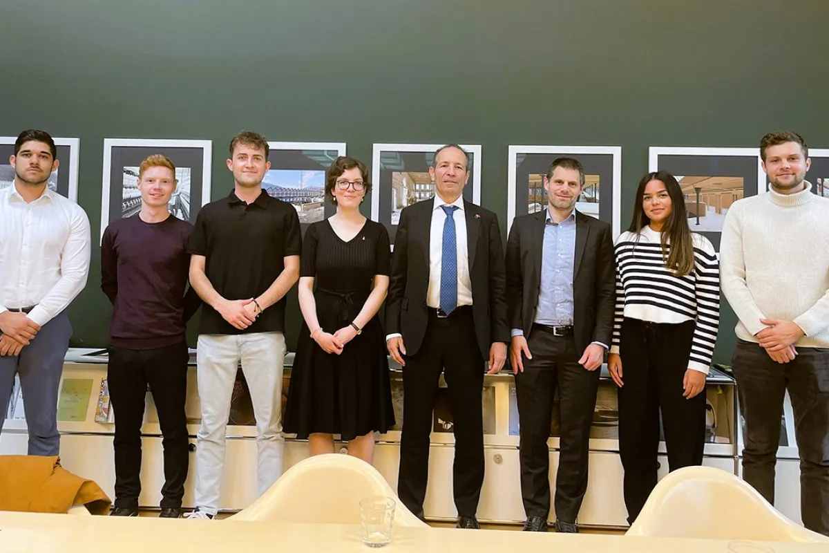 Eine Gruppe von sieben Personen, darunter Studierende und zwei Männer in Anzügen, steht lächelnd vor einer Wand mit Bildern. Sie befinden sich in einem Raum der Universität Liechtenstein.