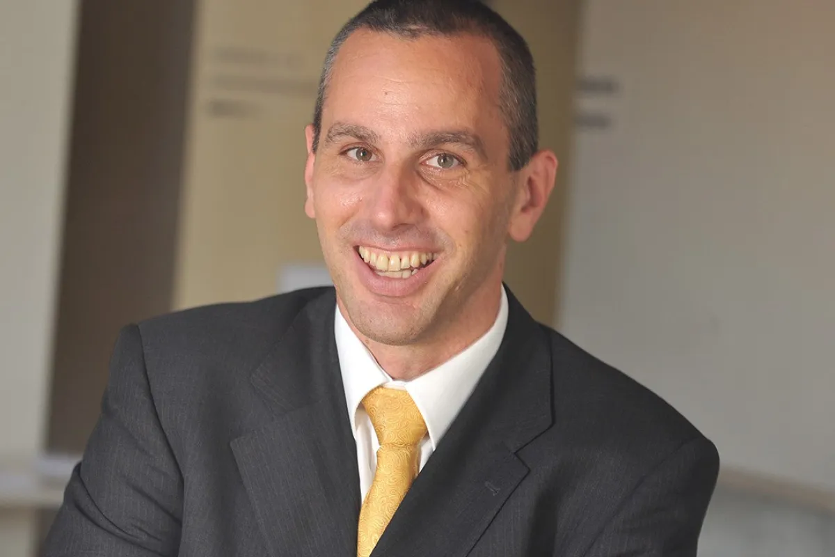 Porträt eines lächelnden Mannes in einem dunklen Anzug mit gelber Krawatte, vor einem neutralen Hintergrund.