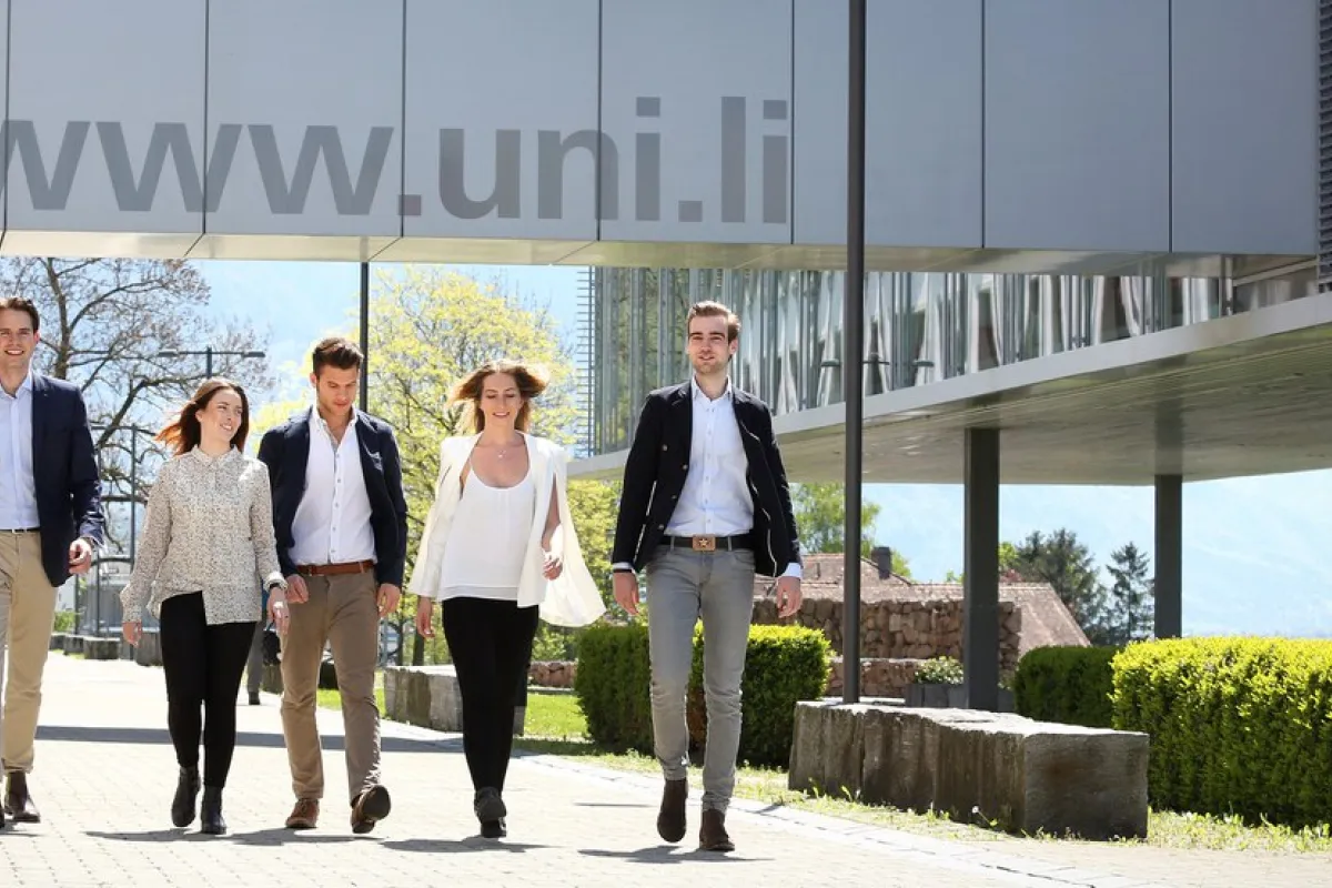Eine Gruppe von fünf Personen geht lächelnd auf einem sonnigen Campusweg der Universität Liechtenstein entlang, im Hintergrund moderne Architektur und Berge.