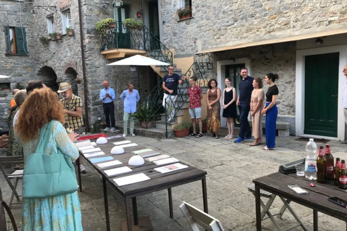 Eine Gruppe von Menschen versammelt sich in einem historischen Innenhof eines Dorfes. Auf Tischen liegen Dokumente und Objekte aus. Die Szene zeigt eine Präsentation oder Diskussion im Freien, umgeben von alten Steinmauern und Pflanzen.