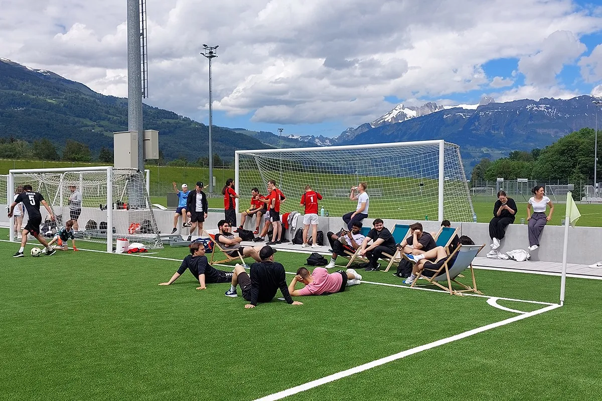 Teilnehmende des Völkerballturniers entspannen sich auf einem Sportplatz vor einer beeindruckenden Bergkulisse. Einige sitzen auf Liegestühlen, während andere am Spielfeldrand stehen. Im Hintergrund sind Fußballtore und Spieler in Sportkleidung zu sehen.