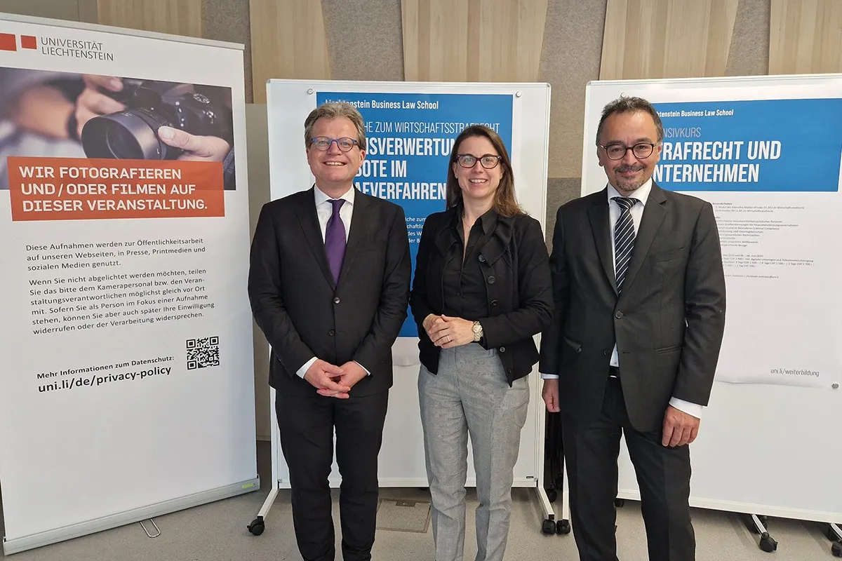 Drei Personen stehen lächelnd vor Informationsplakaten der Universität Liechtenstein. Die Plakate thematisieren Wirtschaftsstrafrecht und Beweisverwertungsverbote. Die Personen tragen formelle Kleidung.