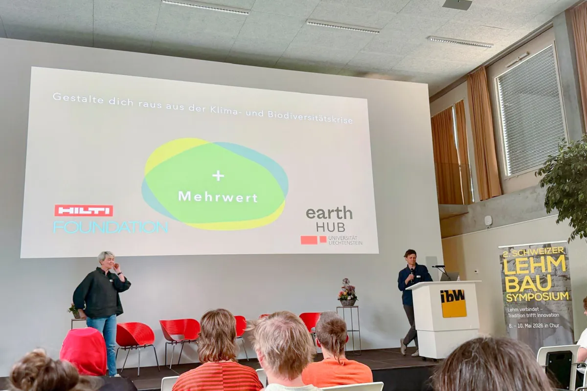 Zwei Vortragende vor einer Leinwand mit orangen Sesseln