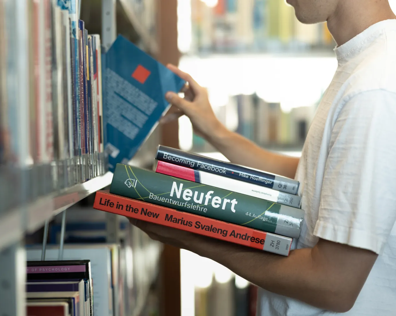 Student wählt Fachliteratur aus dem Bücherregal der Universitätsbibliothek Liechtenstein, mehrere Bücher im Arm.