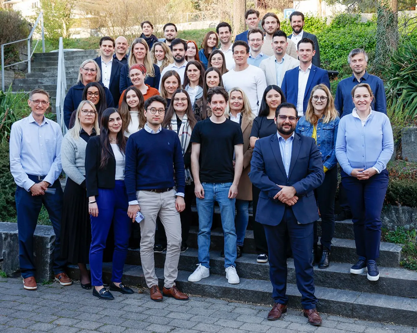 Gruppenfoto von Studierenden des Diplomstudiengangs Treuhandwesen an der Universität Liechtenstein, die auf einer Steintreppe im Freien stehen. Die Gruppe besteht aus Männern und Frauen in formeller Kleidung, umgeben von grüner Vegetation.