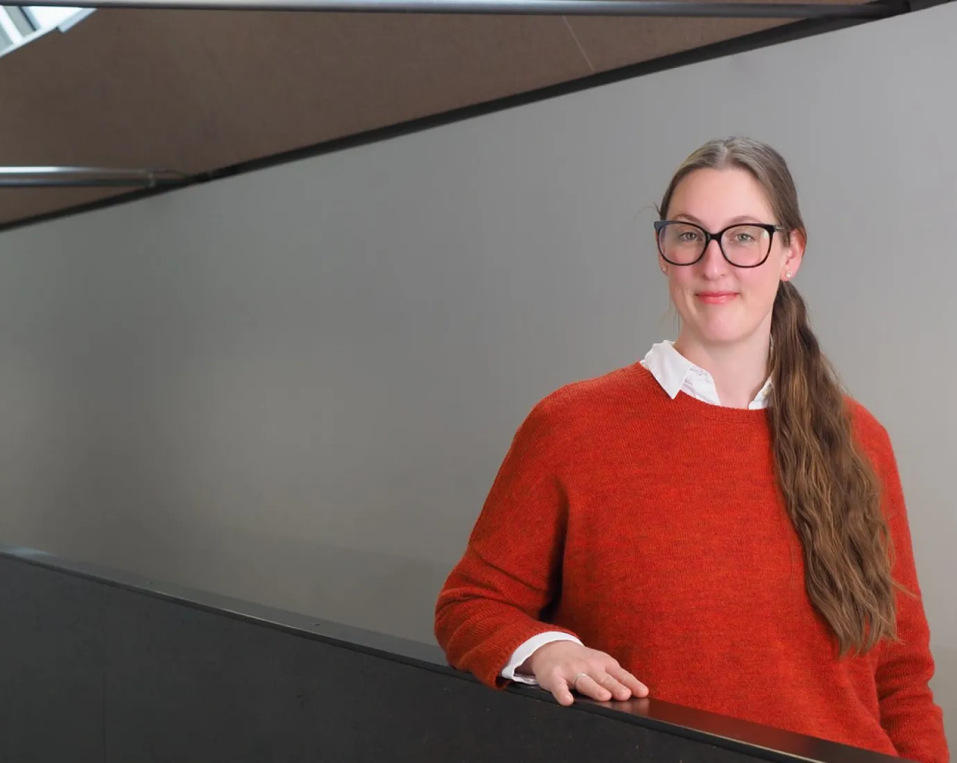 Eine Person mit Brille und langem Haar steht in einem modernen Gebäude und trägt einen roten Pullover. Sie lächelt leicht und lehnt sich an ein Geländer.