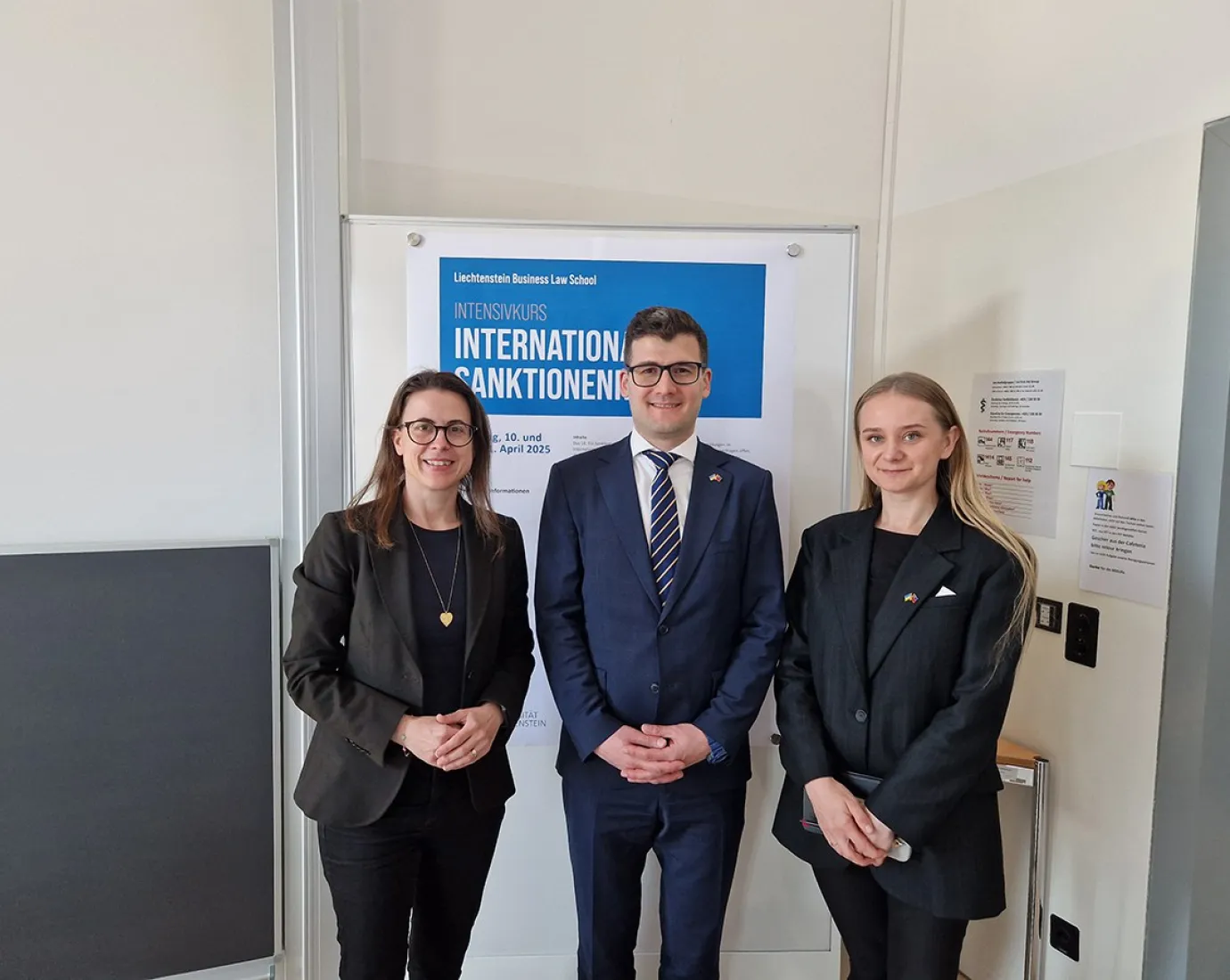 Drei Personen stehen vor einem Poster mit der Aufschrift "Intensivkurs Internationales Sanktionenrecht". Sie tragen formelle Kleidung und lächeln in die Kamera.