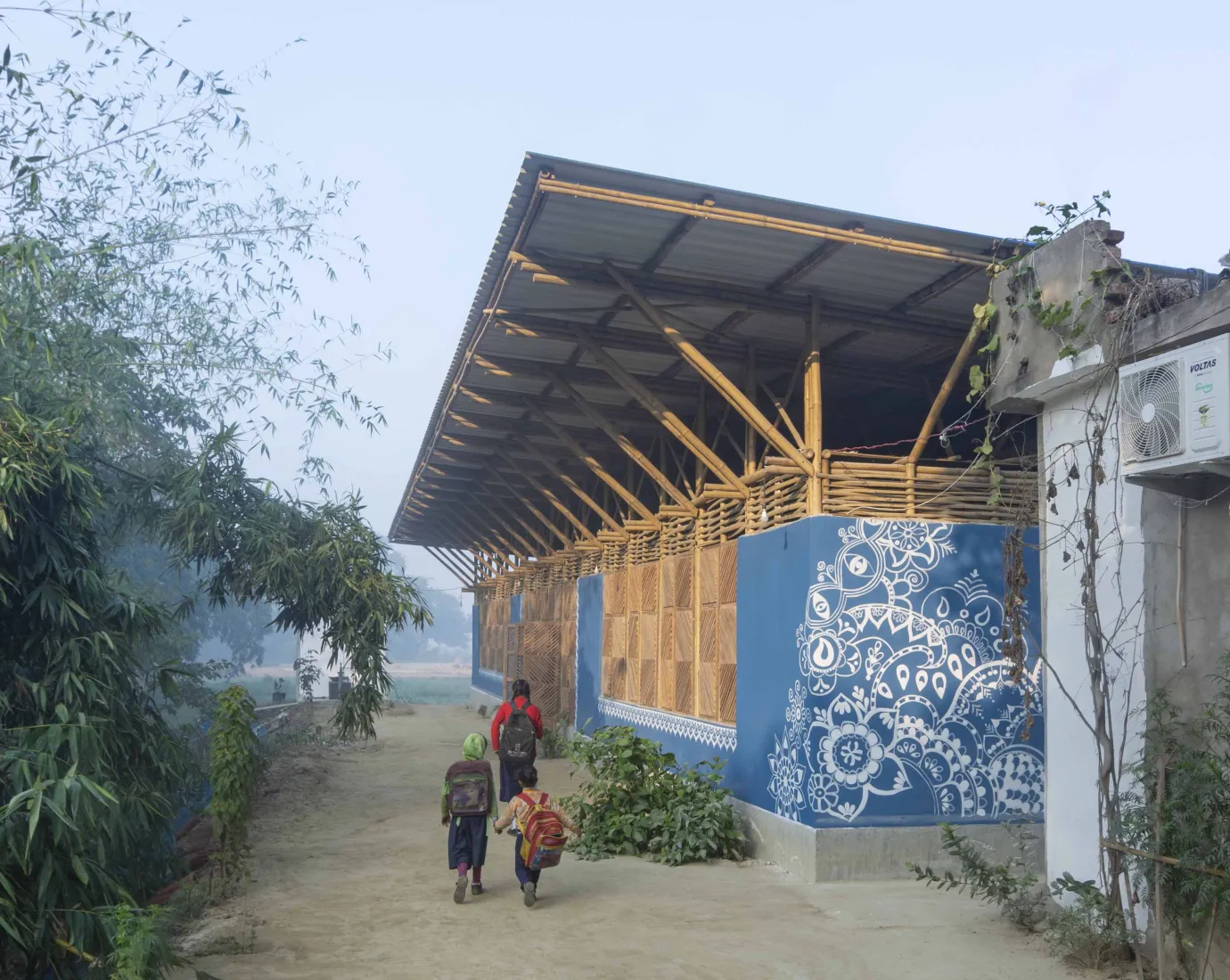 Kinder gehen auf einem sandigen Weg an einem blau bemalten Bambusgebäude mit weißem Ornamentmuster vorbei.