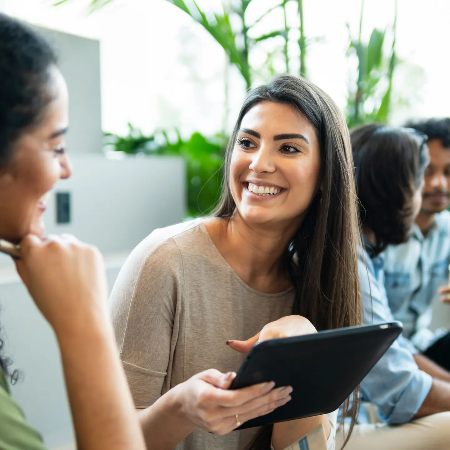  Eine Gruppe junger Menschen sitzt an einem Tisch und unterhält sich lebhaft. Die Stimmung ist fröhlich und positiv. Im Fokus des Bildes steht eine Frau, die ein Tablet in der Hand hält.
