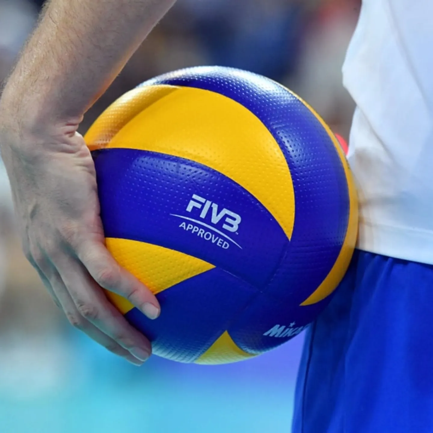 Person, die vor einem unscharfen Hintergrund einen blau-gelben Volleyball mit der Aufschrift „FIVB Approved“ hält.