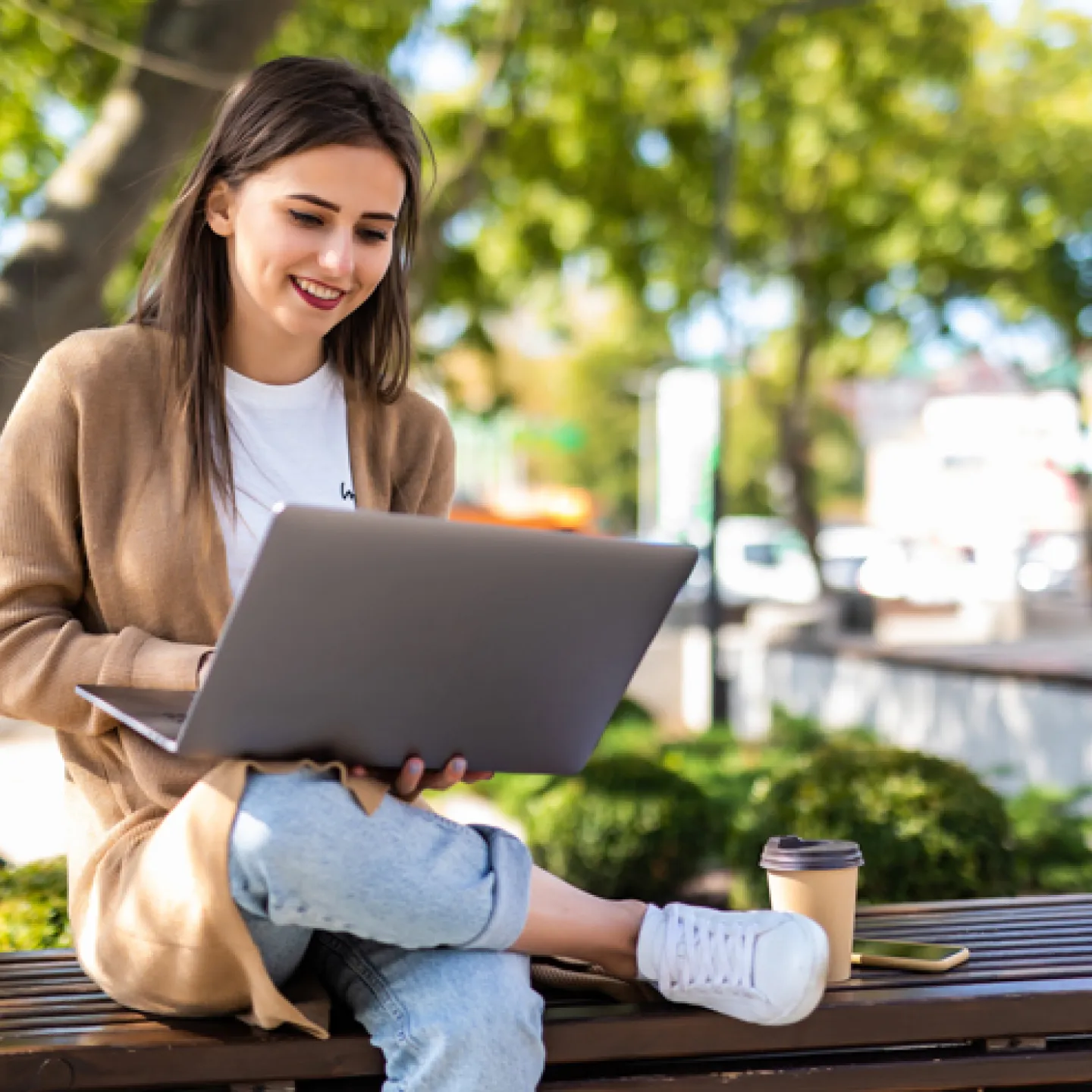 Eine junge Frau sitzt lächelnd auf einer Parkbank im Freien und arbeitet an ihrem Laptop. Sie trägt eine beige Strickjacke, ein weißes T-Shirt und Jeans. Neben ihr auf der Bank stehen ein Kaffeebecher und ihr Smartphone. Im Hintergrund sind grüne Bäume und unscharfe städtische Elemente zu sehen, die eine entspannte und produktive Atmosphäre vermitteln.