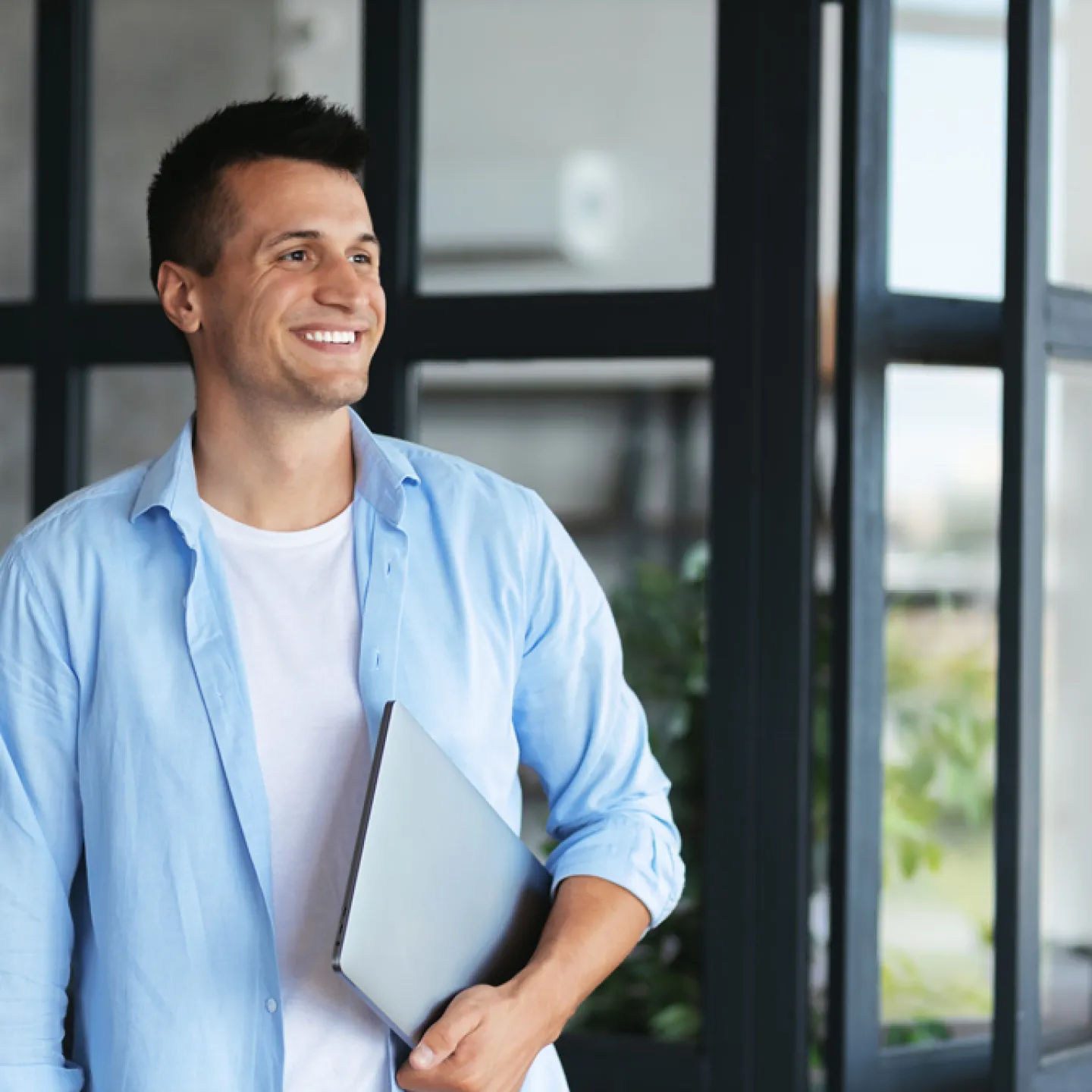 Ein junger Mann lächelt und blickt zur Seite, während er einen Laptop unter dem Arm hält. Er trägt ein hellblaues Hemd über einem weißen T-Shirt und steht in einem modernen Innenraum mit Glaswänden und Pflanzen im Hintergrund. Die Atmosphäre wirkt entspannt und professionell.