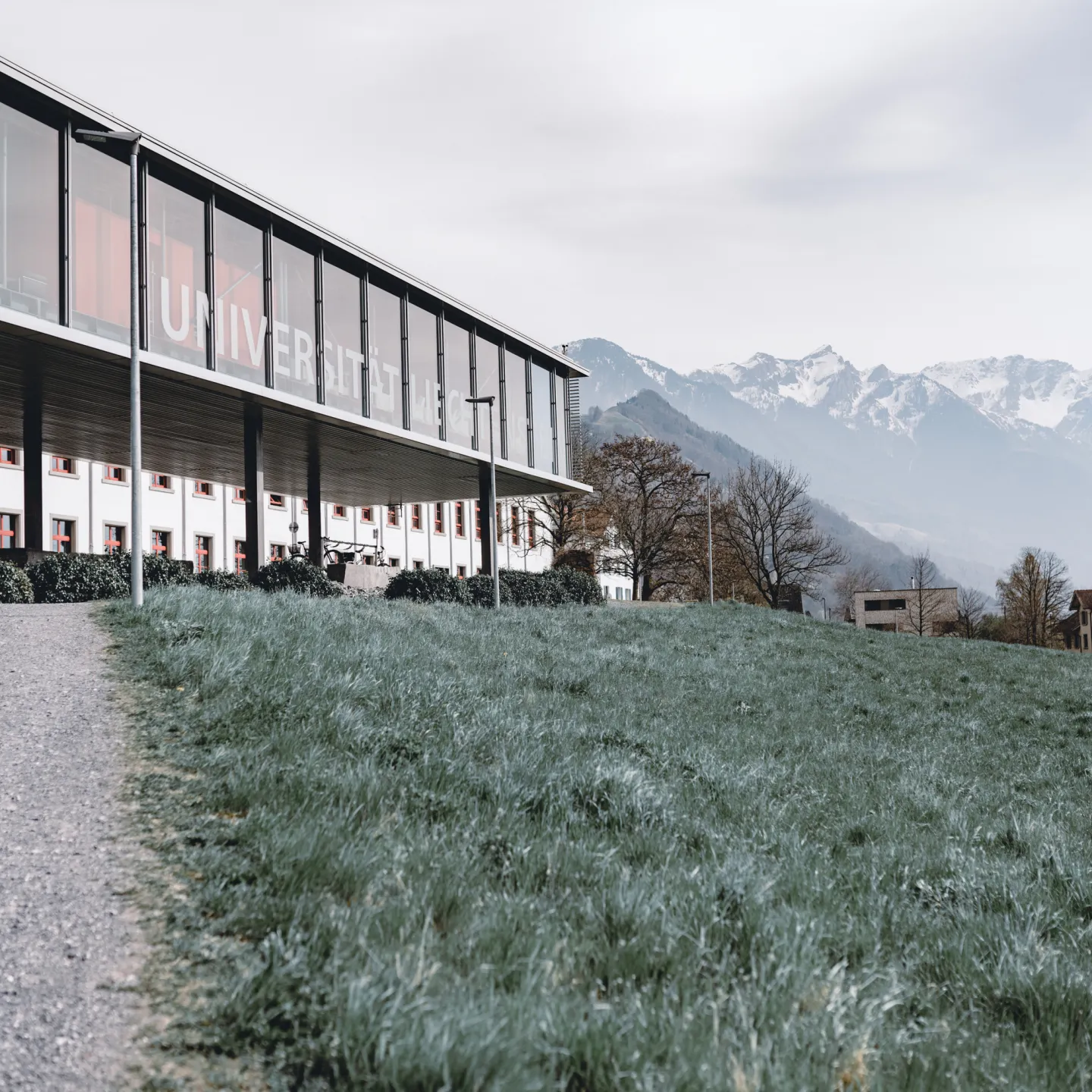 Campus Liechtenstein mit Glasfassade, Wiesenweg und Panoramablick auf die umliegenden Berge und das Dorf Vaduz.