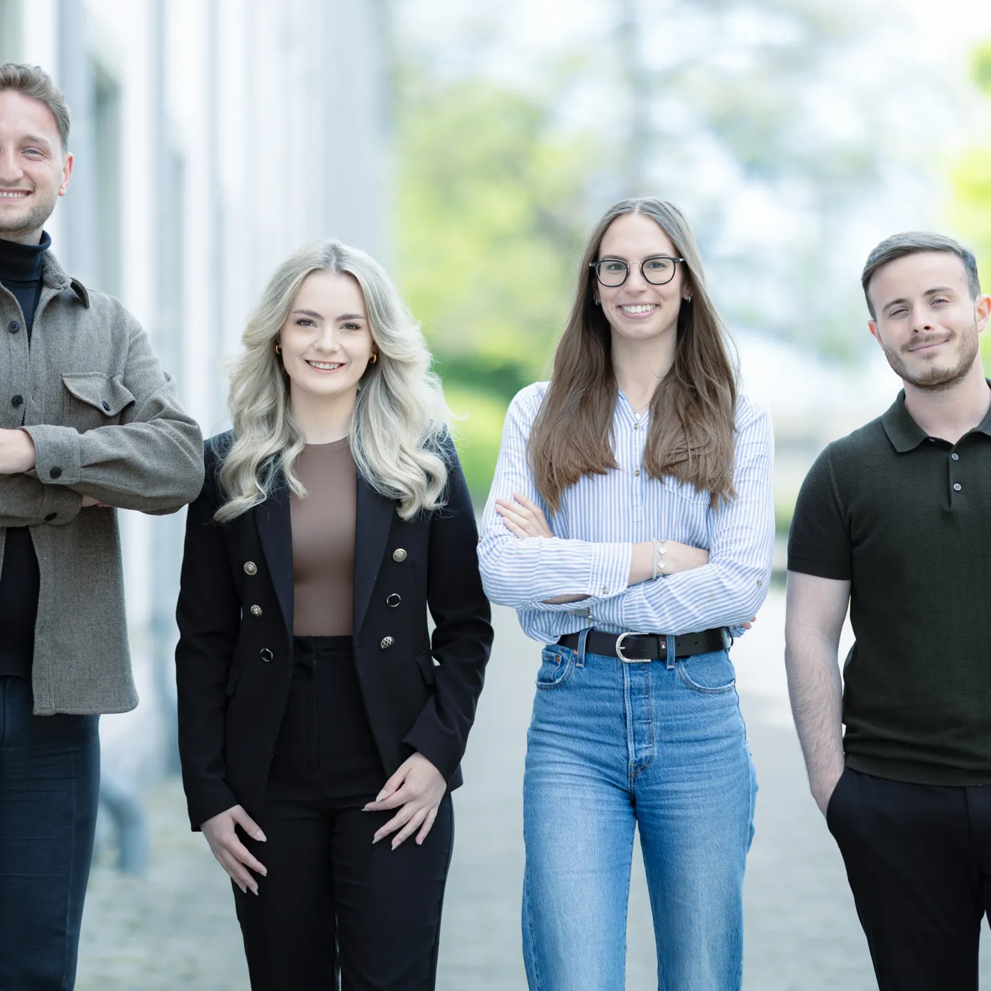 Vier Studierende im Masterstudiengang Information Systems der Universität Liechtenstein stehen nebeneinander auf einem Gehweg vor einem Universitätsgebäude und lächeln in die Kamera. Die Kleidung reicht von Business- bis Casual-Stil.