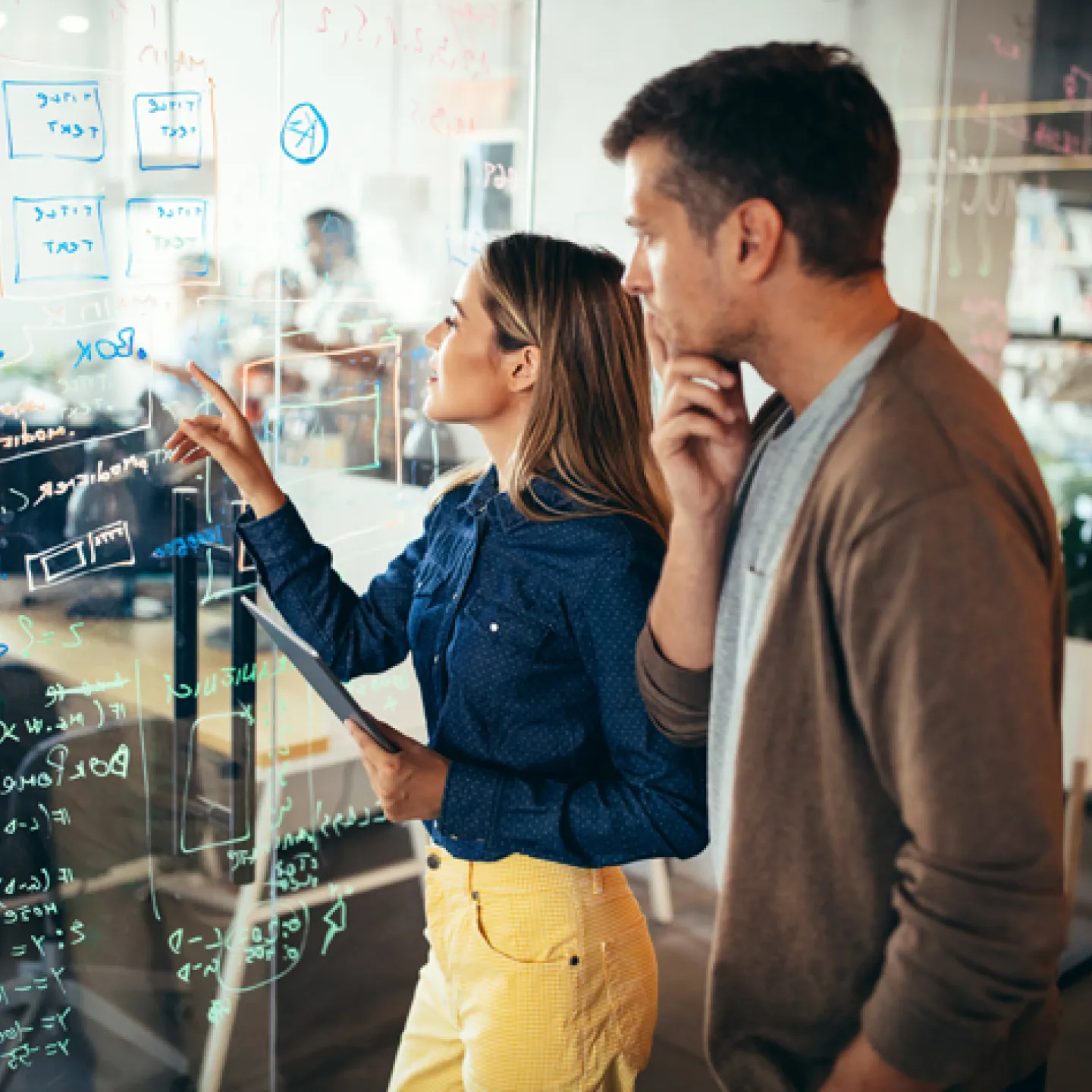 Ein Mann und eine Frau betrachten gemeinsam eine große Glaswand, die mit Notizen und Diagrammen beschrieben ist. Beide wirken nachdenklich und konzentriert, während sie an einer komplexen Aufgabe arbeiten.