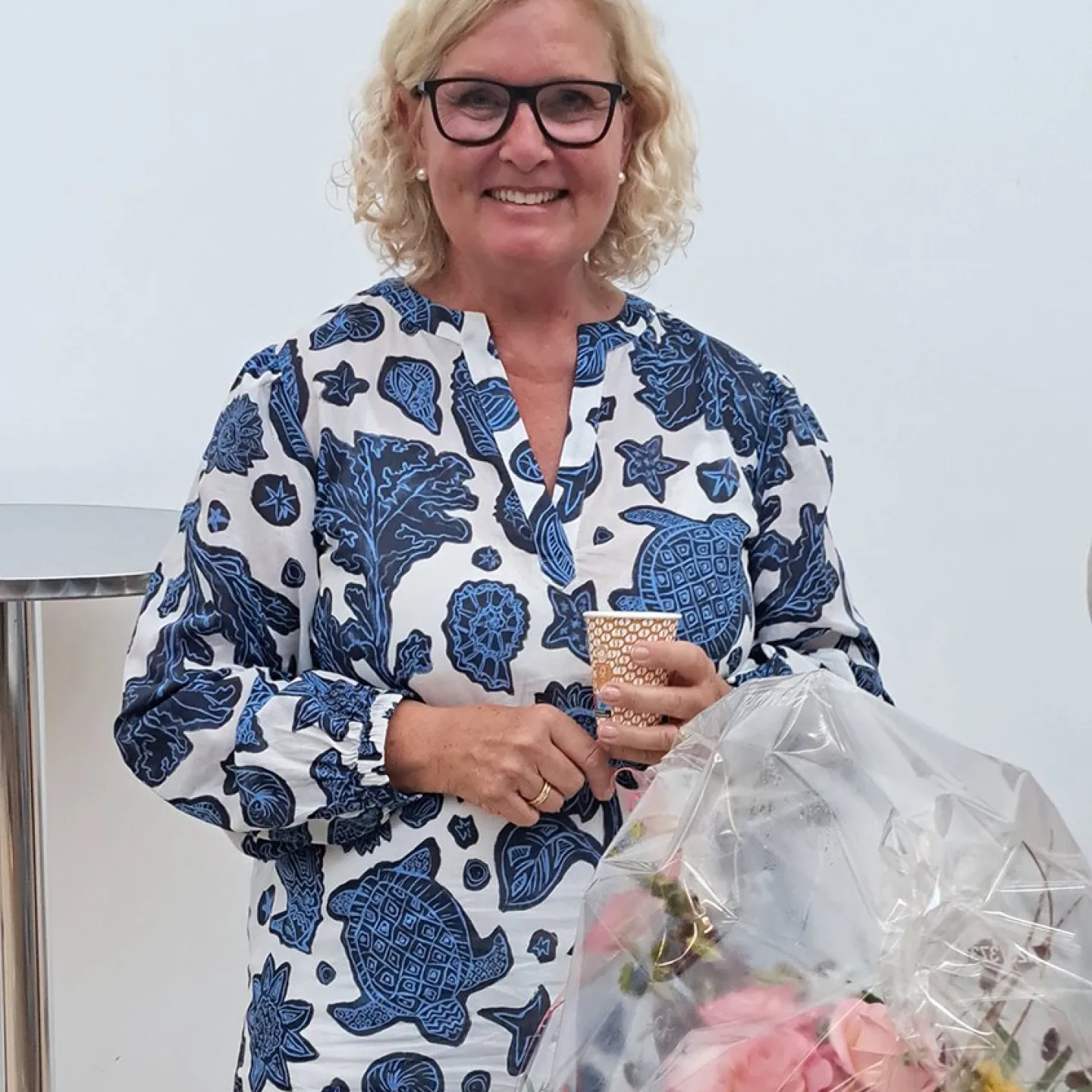 Eine lächelnde Frau mit blonden Haaren und Brille hält einen Becher in der Hand. Sie trägt ein blau-weiß gemustertes Kleid. Vor ihr steht ein Blumenstrauß in Folie verpackt.