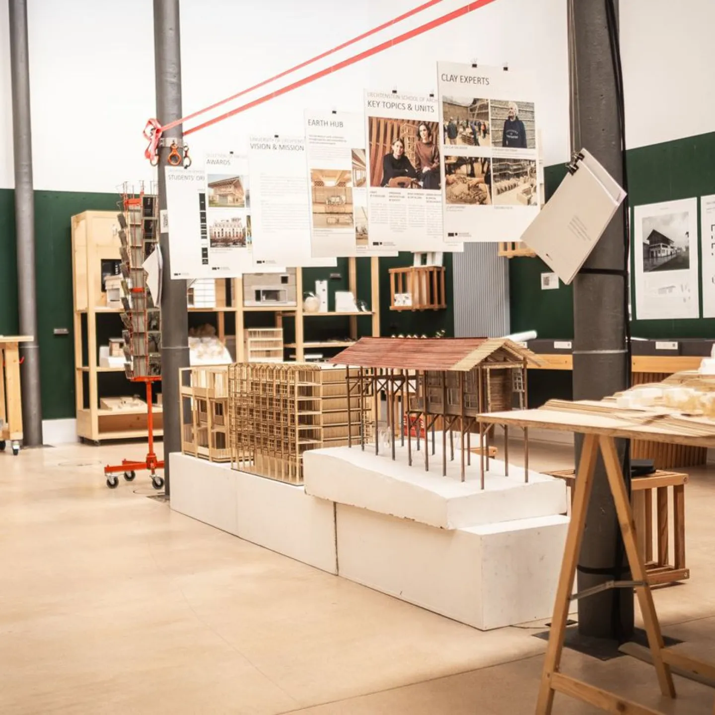 Architektur-Studienraum mit Modellen und Plakaten. Verschiedene Architekturmodelle aus Holz und Papier sind auf Tischen ausgestellt. An den Wänden hängen Plakate mit Informationen zu Projekten und Themen. Der Raum ist hell und modern gestaltet.