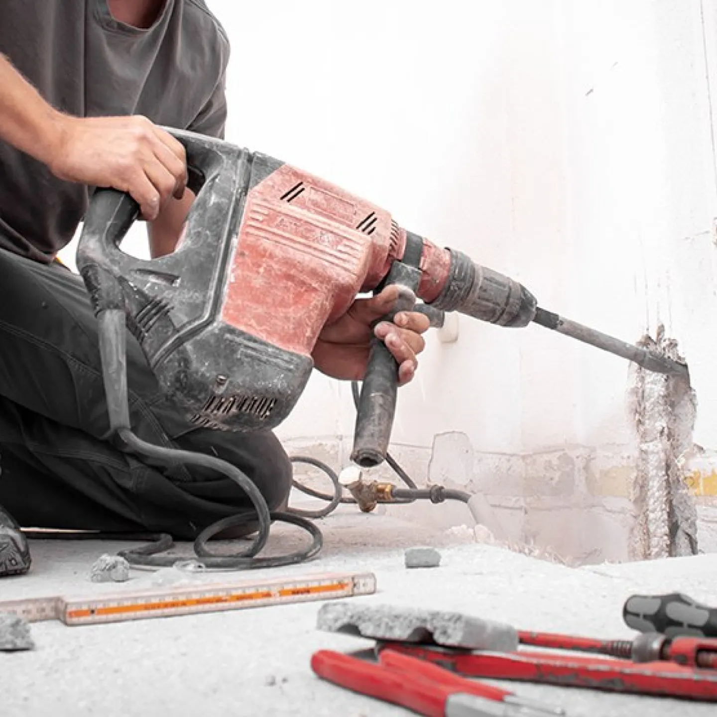 Ein Bauarbeiter kniet auf dem Boden und benutzt einen roten Kombi-Hammer, um eine Wand zu bearbeiten. Im Vordergrund liegen Werkzeuge und Bauutensilien.