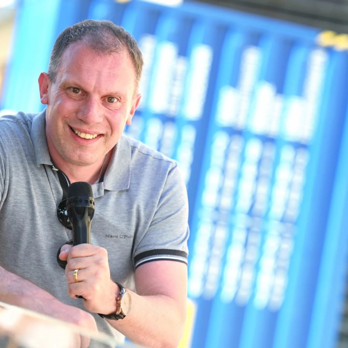 Andreas Kräätli lächelnd in einem grauen Polohemd hält ein Mikrofon. Im Hintergrund sind verschwommene blaue Strukturen zu sehen.