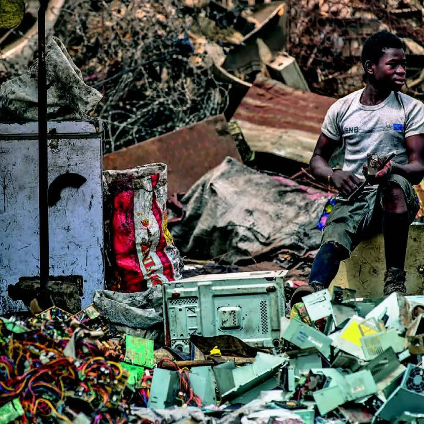 Ein Mann sitzt auf einem Hocker inmitten von Elektroschrott auf einer Müllhalde. Um ihn herum sind alte Geräte und Kabel zu sehen. Die Szene zeigt die Problematik von Elektroschrott und dessen Auswirkungen auf die Umwelt.