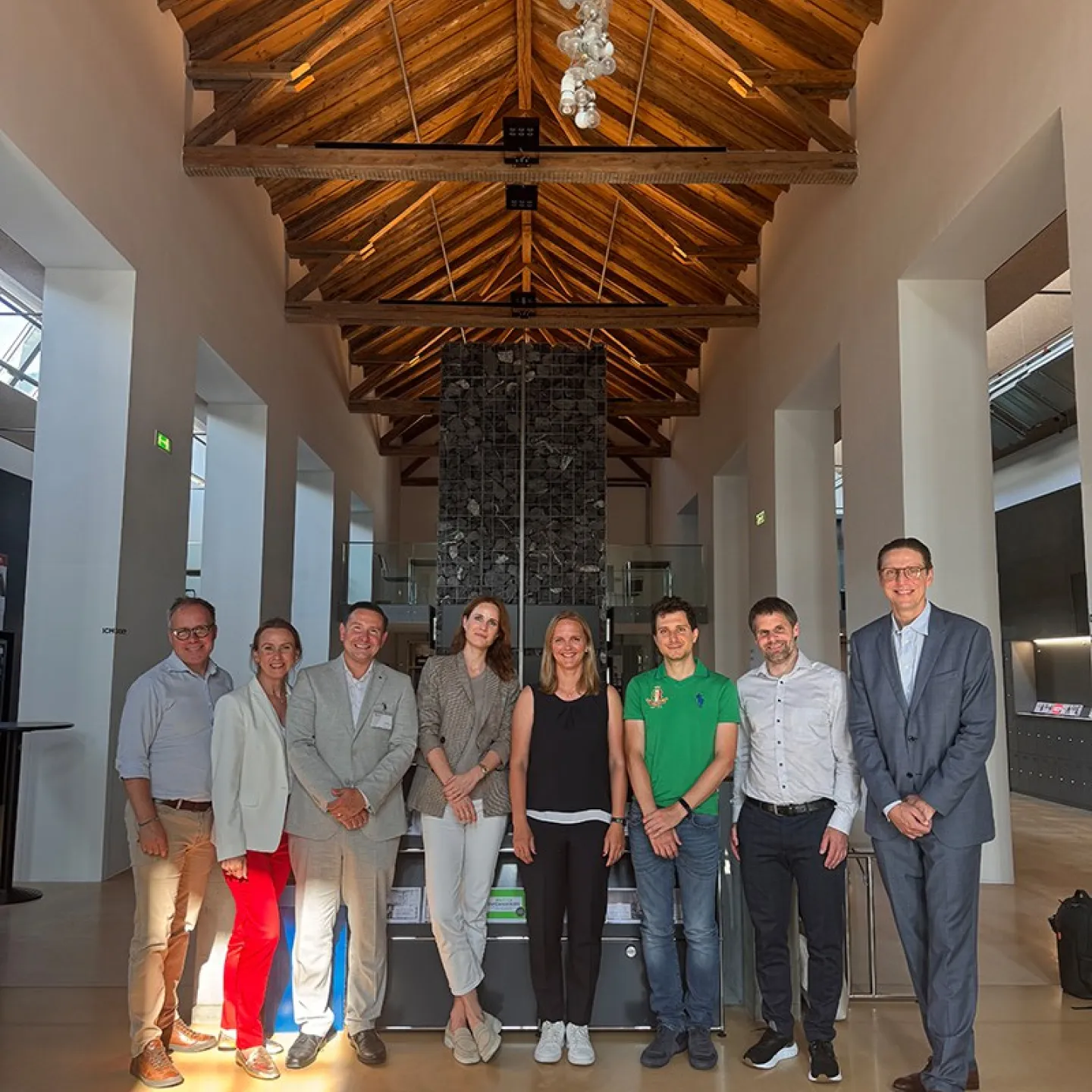 Eine Gruppe von neun Personen steht in einer modernen Halle mit Holzdach und einem Kronleuchter. Sie lächeln in die Kamera. Im Hintergrund sind hohe Fenster und eine Steinwand zu sehen.