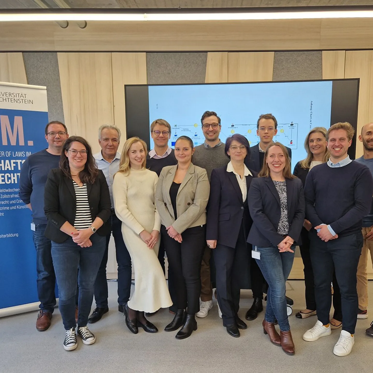 Gruppenfoto von Studierenden und Dozenten des Executive Masters of Law im Wirtschaftsstrafrecht an der Universität Liechtenstein. Im Hintergrund ein Bildschirm mit Präsentationsfolie und ein Roll-up mit Informationen zum Studiengang.