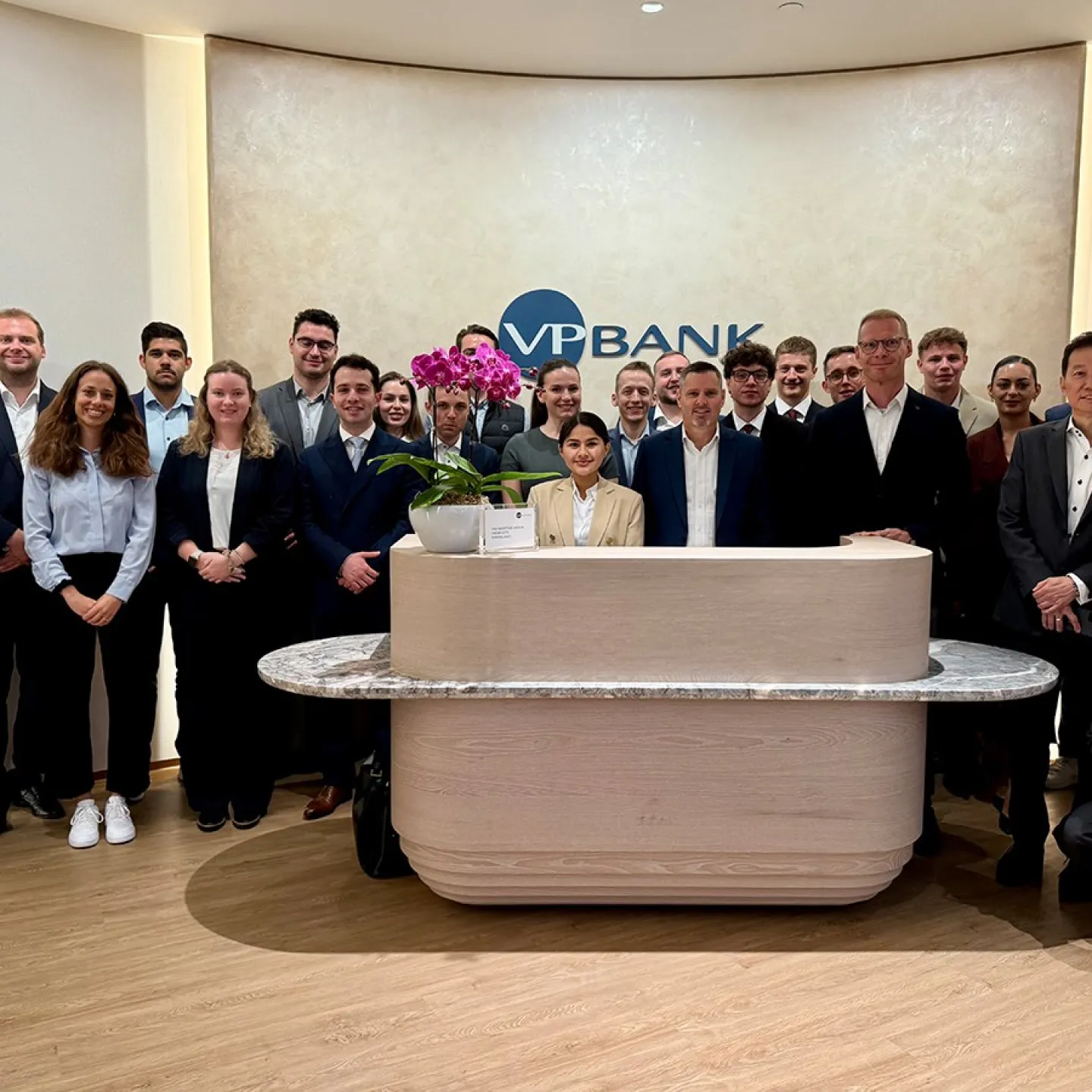 Gruppe von Studierenden und Fachleuten in formeller Kleidung steht in der Lobby der VP Bank. Im Hintergrund ist das Logo der Bank sichtbar. Die Szene vermittelt eine professionelle und lernorientierte Atmosphäre.