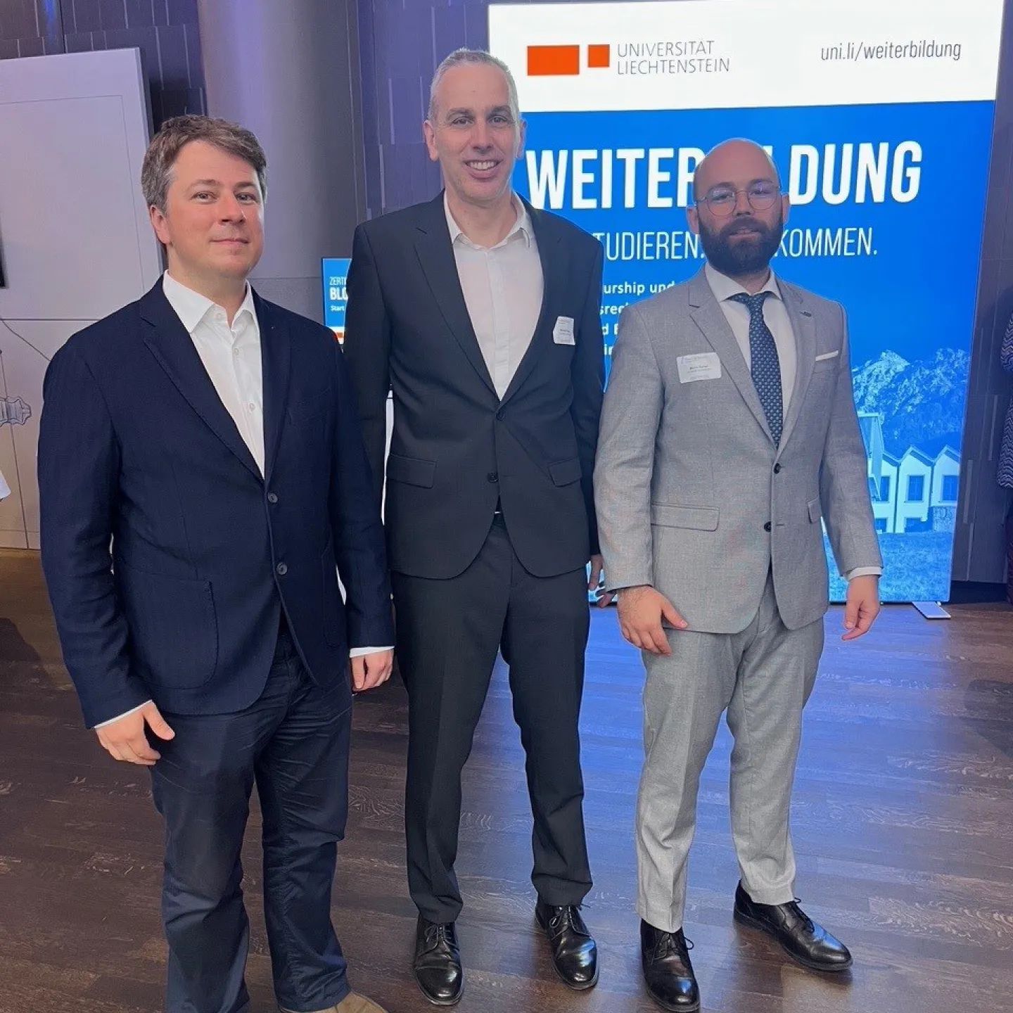 Drei Männer in Anzügen stehen vor einem Schild der Universität Liechtenstein. Im Hintergrund ist ein Banner mit der Aufschrift "Weiterbildung" zu sehen.